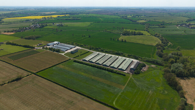 Aerial view of large agricultural farm buildings and industrial sheds surrounded by lush green fields and hedgerows in the rural landscape of Kettering, England, United Kingdom.