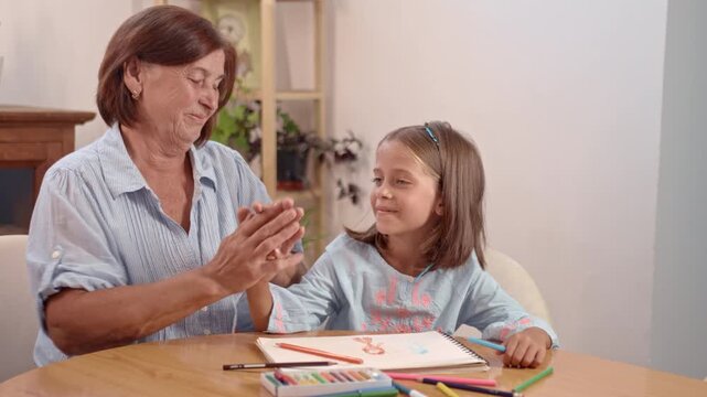 A grandmother helps her granddaughter with art using crayons and a sketchbook at a table. They share a moment of fun and creativity in their home.