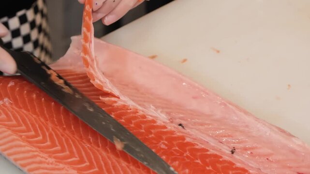 A close-up shot capturing the precise technique of a professional chef using a long filleting knife to slice skin and bone from a vibrant, fresh raw salmon fillet in a commercial kitchen environment. 