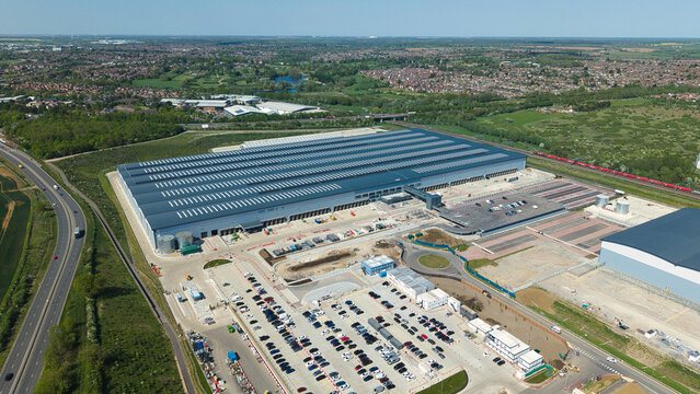 Aerial view of the large industrial warehouse at Symmetry Park with parking lots and surrounding green landscape under a clear blue sky in Kettering, England, United Kingdom.