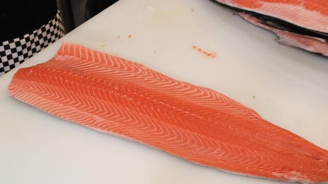 A close-up, high-angle shot of a chef's hands skillfully using a sharp filleting knife to slice a vibrant, fresh raw salmon fillet. The footage showcases the rich orange texture and natural fat marbli