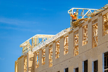 Construction worker in safety gear installing wooden roof trusses on a large multi-story building framing against a clear blue sky, USA © Andrei Liashchou