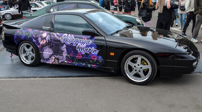 Black Nissan 180SX with custom itasha anime graphics stands at outdoor car meet. Sleek coupe features deep dish rims and Kitagawa Marin decals amidst crowd of enthusiasts
