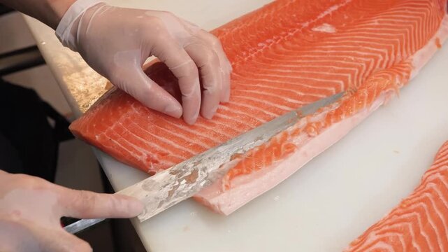 Close-up of a professional chef's hands in gloves skillfully using a sharp fillet knife to cut and prepare a large, vibrant orange raw salmon fillet on a white industrial cutting board. The footage hi
