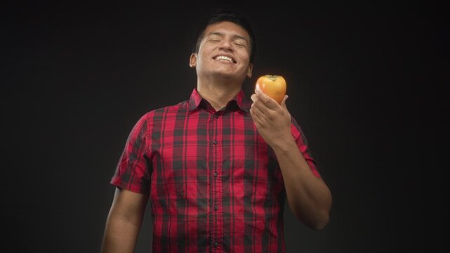 Young man holding ripe persimmon fruit in right hand, smiling and presenting it while wearing red plaid shirt in a dark studio setting; joyful.