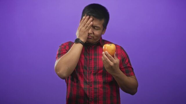 Man in red plaid shirt holding a persimmon and covering his face with his hand in a studio purple backdrop; disgust hesitation.