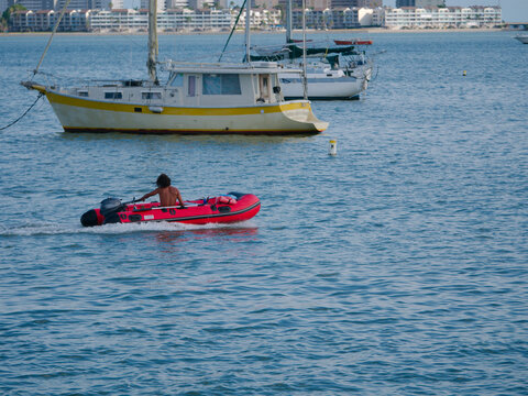 A man steers a red inflatable motorboat in Gulfport, FL across calm blue harbor waters past moored yachts and coastline buildings, evoking summer leisure, coastal travel and nautical lifestyle in an u