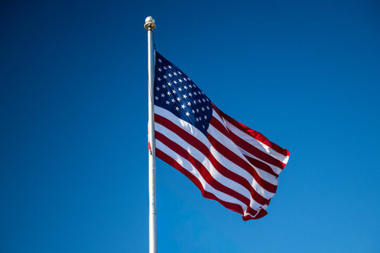 A red, white, and blue colored American flag hanging on a metal flagpole. There are blue skies and clouds in the background. There's no wind, and the flag is drooping downward from the gold pole. 