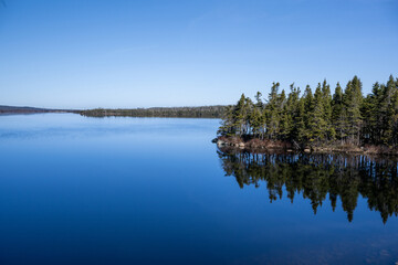 A large, wide, deep blue lake with a calm, smooth surface. There are tall, mature, evergreen trees along the rocky shoreline, reflecting in the freshwater.  The surface is like a mirror, reflecting. © Dolores  Harvey
