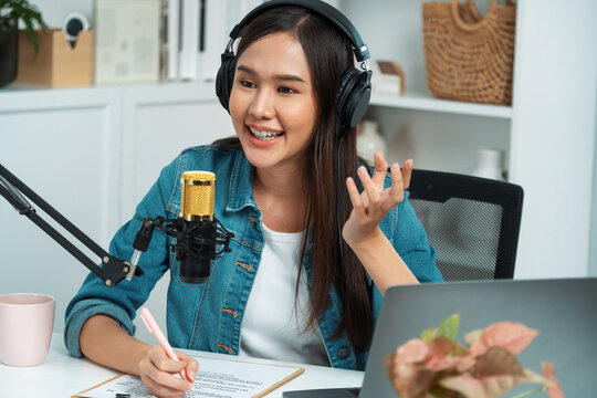 Host channel Asian influencer talking in broadcast streaming online wearing headset on social media live with script note, greeting listeners in life coaching or marketing at modern studio. Stratagem.