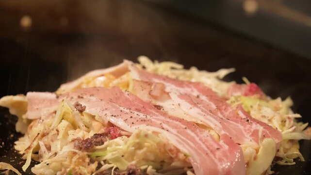 Close-up footage of traditional Hiroshima-style okonomiyaki being prepared on a professional iron griddle. The scene highlights the layered construction of the savory pancake, featuring a generous amo