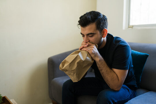 Stressed man breathing into a paper bag, experiencing a panic or anxiety attack at home, mental health concept
