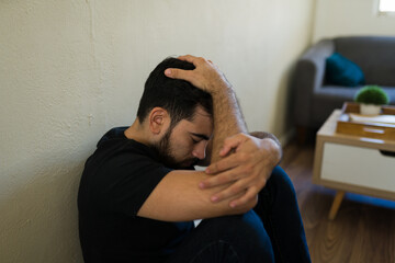 Obraz premium Depressed man experiencing sadness and anxiety, sitting alone on the floor with head in hands, struggling with mental health issues