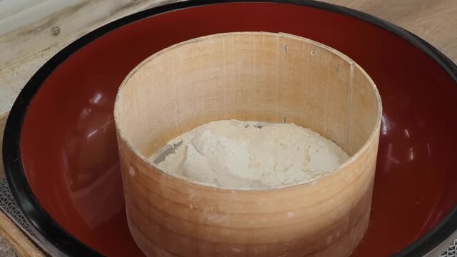 Close-up footage of the traditional Japanese process of making handmade soba noodles. Fine buckwheat flour is sifted through a round wooden sieve into a large, deep-red lacquer kneading bowl (konebach