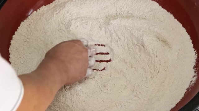 High-angle, close-up footage of a person&rsquo;s hand mixing dry whole wheat flour (atta) in a large red plastic bowl. The video captures the texture of the finely ground flour being swirled and tossed, a k