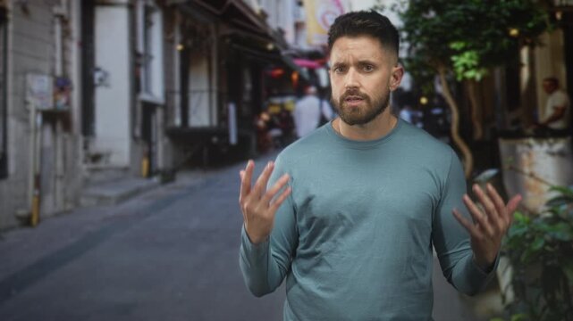 Man with beard raises hands palms up in a questioning shrug on a narrow street lined with buildings and plants; confusion.