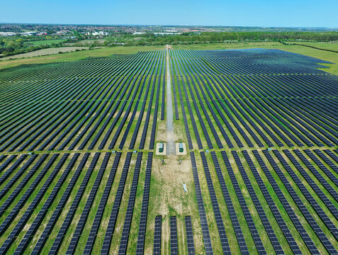 Aerial view of a large solar farm with rows of photovoltaic panels in green fields under a clear blue sky in Little Irchester, England, United Kingdom.