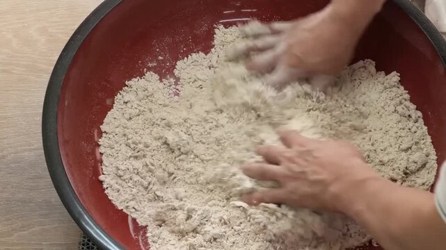 close-up shot showing an artisan's hands skillfully mixing and kneading fresh buckwheat flour with water in a traditional large red and black lacquer bowl. This authentic Japanese culinary process, kn