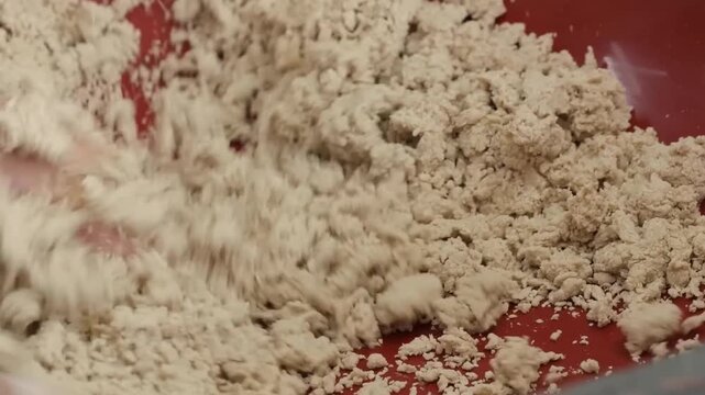 High-angle, close-up footage of a man's hands traditionally kneading whole wheat flour (atta) with water in a large red bowl. The mixture is in the early stages of clumping together to form a soft dou