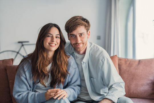 Smiling young couple sit close on a cozy couch in a bright living room, sharing a relaxed moment together that conveys warmth, intimacy, friendship and modern home lifestyle.