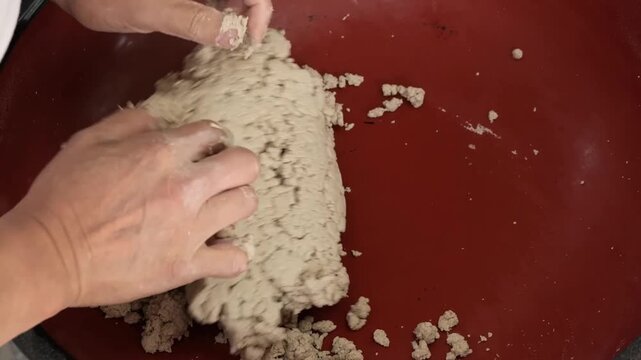 A close-up footage shot showing a pair of hands actively mixing and crumbling a light-brown, textured material that resembles raw dough, starch, or specialized construction material like fine river sa