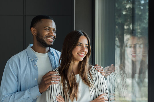 Smiling couple stands by a window at home, gazing outside and sharing a quiet, optimistic moment. Natural light and relaxed body language convey affection, trust and plans for the future together.