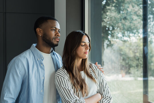 A thoughtful young couple share a quiet moment of emotional support and comfort as they stand by a large window, conveying intimacy, contemplation and togetherness in a calm modern home.