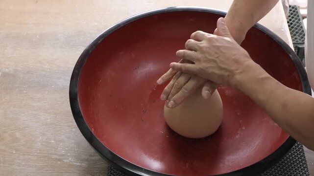A close-up shot of skilled hands meticulously kneading and shaping a smooth, teardrop-shaped mass of dough or clay inside a traditional large red and black lacquer mixing bowl. The footage captures th