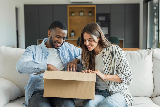 A smiling couple sits on a living room sofa opening a cardboard box together, sharing a joyful moment while unpacking a delivery in their modern, cozy home.