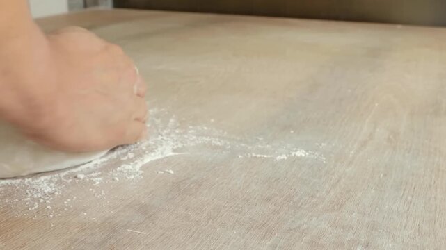 A close-up shot of a baker's hand delicately sprinkling white wheat flour over a smooth, round lump of fresh pizza or bread dough resting on a rustic wooden tabletop in a kitchen.