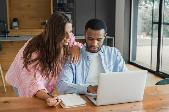 A young couple sits at a wooden table in a bright kitchen, focused on a laptop and notebook as they plan finances, schedule, and household tasks while collaborating calmly and supportively.