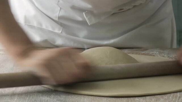 High-angle, close-up shot of a professional chef using a wooden rolling pin to flatten a ball of fresh dough. The footage shows the rhythmic motion of shaping the dough into a smooth, thin circle on a