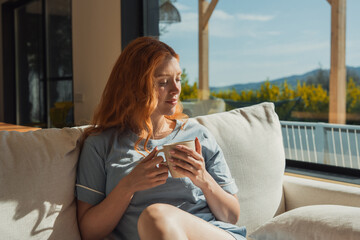 Obraz premium A red-haired woman sits on a light sofa holding a mug, enjoying warm morning sunlight through large windows onto a terrace and distant hills, conveying a calm, cozy and reflective at-home moment.