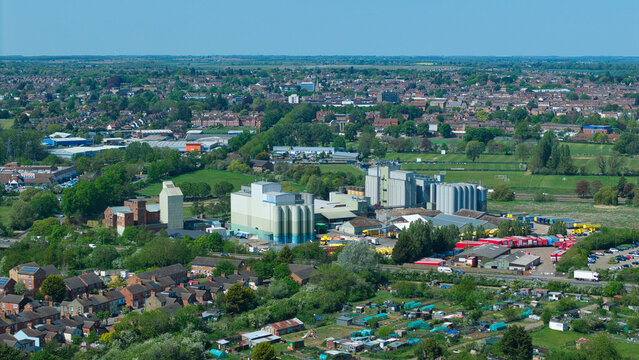 Aerial view of large industrial silos and warehouses surrounded by residential houses, green trees, and allotments in Little Irchester, England, United Kingdom.