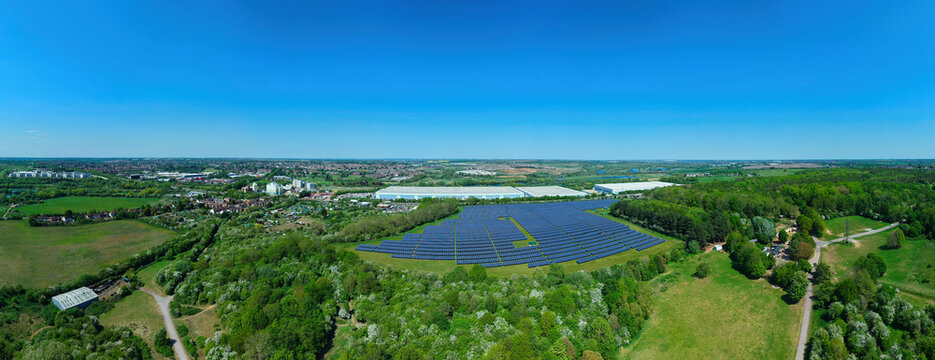 Aerial view of a large solar farm with rows of blue panels nestled among lush green trees and fields under a clear sky near Little Irchester, England, United Kingdom.