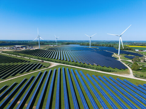 Aerial view of Chelveston Wind Farm featuring rows of solar panels and tall wind turbines under a clear blue sky in the rural landscape of Wellingborough, England, United Kingdom.