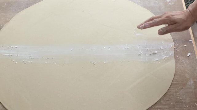 A top-down, close-up shot of a chef's hand skillfully sprinkling white flour across a large, perfectly circular piece of rolled-out dough. The dough is laid out on a wooden workstation, and the moveme