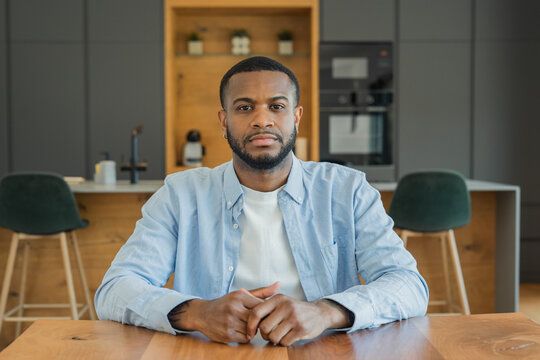 Young man sits at a wooden kitchen table in a modern home, looking directly at the camera with a thoughtful, calm expression. Natural light and casual clothing create a comfortable lifestyle and remot