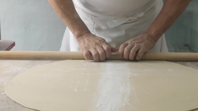 High-angle close-up of a professional baker's hands skillfully rolling out a thin sheet of fresh pastry dough using a traditional long wooden rolling pin. The scene captures the authentic process of p