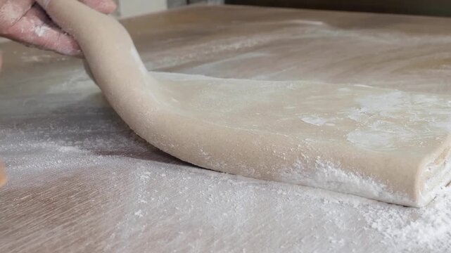 Close-up shot of a baker's hands skillfully folding layers of fresh pastry dough on a wooden workstation. The surface is heavily dusted with white flour, highlighting the traditional process of making