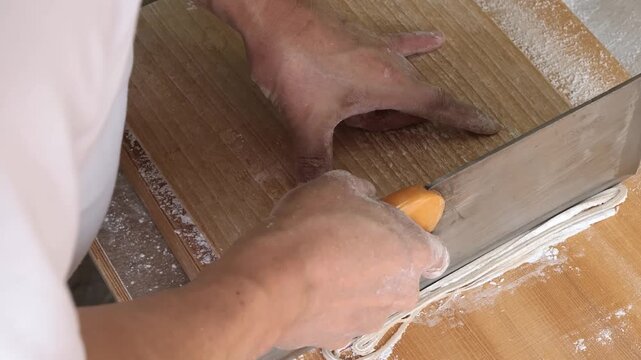 A detailed, high-angle close-up shot of a professional chef using a traditional Japanese soba-kiri (large rectangular knife) and a wooden guide board (komaita) to hand-cut fresh buckwheat dough into t