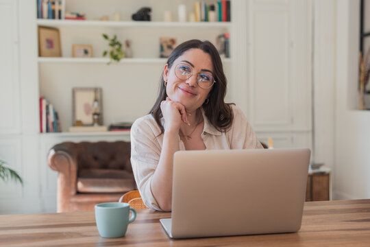 A smiling young woman works on her laptop in a bright home office, looking relaxed and thoughtful as she enjoys a coffee mug and casual, cozy surroundings while working remotely.