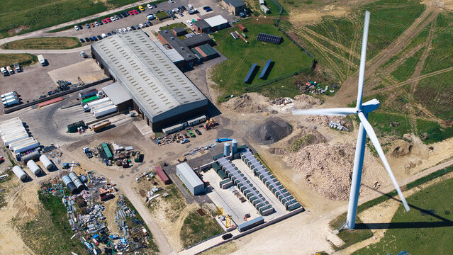 Aerial view of Chelveston Wind Farm featuring a large wind turbine next to an industrial warehouse and parking lot in Wellingborough, England, United Kingdom.