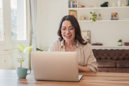 Smiling woman using a laptop for a video call in a bright, cozy home office, engaged in remote work or an online meeting, conveying warmth, focus and connection.