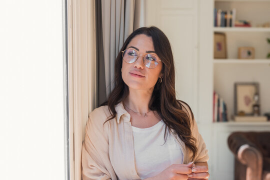 A thoughtful young woman in glasses gazes out a window in a bright, cozy living room, wearing casual clothing and a soft smile that conveys calm, reflection and quiet confidence.