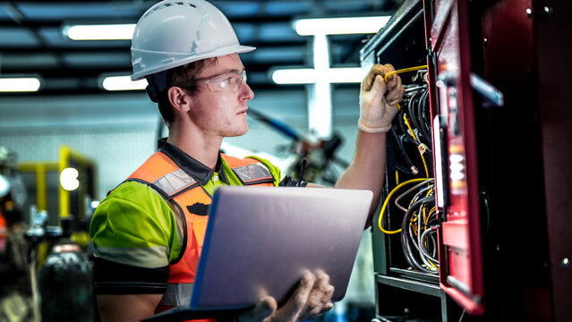 A male engineer in a hard hat holds a laptop while inspecting electrical wires. He is a specialist doing maintenance on a machine in a high-tech smart factory.