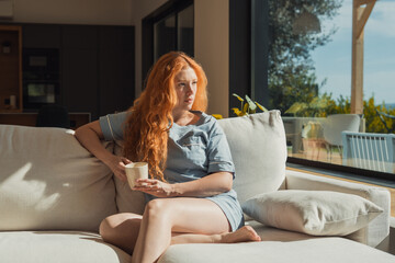 Obraz premium Redhead woman in casual loungewear sits barefoot on a light sofa, holding a mug and gazing out a large window toward a sunny terrace. Warm natural light and calm, reflective mood fill the modern inter