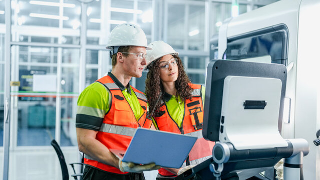 Engineers use a laptop to program an AI system for an industrial robot. The team is in a smart factory cleanroom, working on a high-tech microchip manufacturing line.