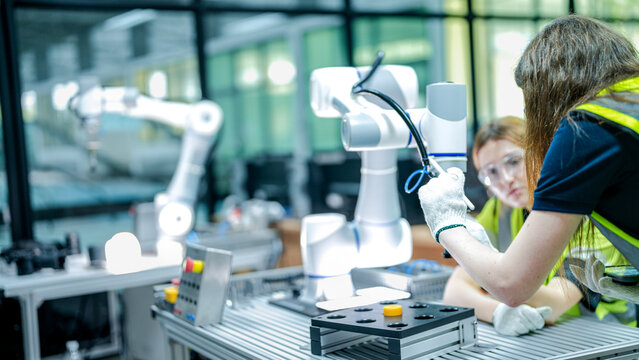 Two female robotics engineers in an R&D lab troubleshoot an AI cobot. One inspects the arm's hardware while the other reviews diagnostic data for the system.
