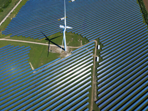 Aerial view of Chelveston Wind Farm featuring a tall wind turbine casting a long shadow over rows of blue solar panels on green fields in Wellingborough, England, United Kingdom.